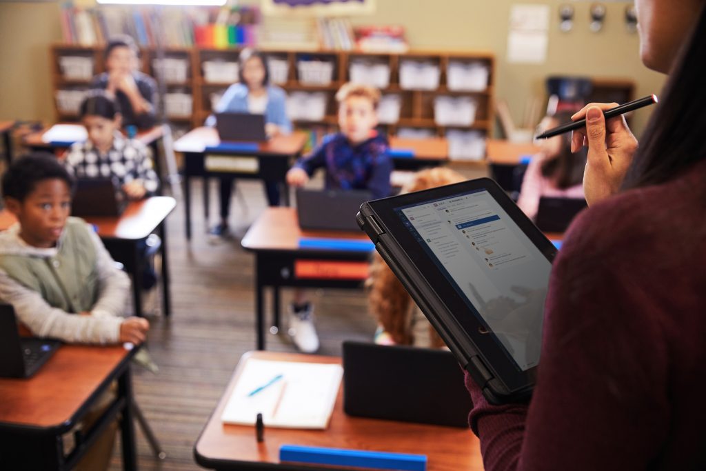 Teacher holding a tablet in a classroom filled with students.