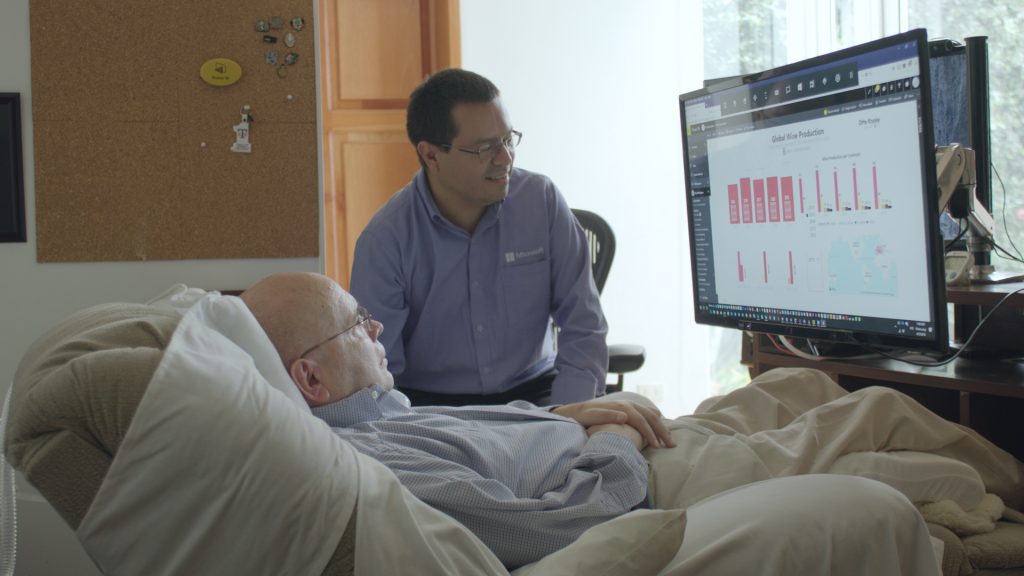 Older man (Otto Knoke) in a hospital bed and younger man (his friend and former colleague, Juan Alvarado) sitting beside him a Power BI report Knoke created using eye-tracking software for Windows 10 on a display monitor at the foot of the bed