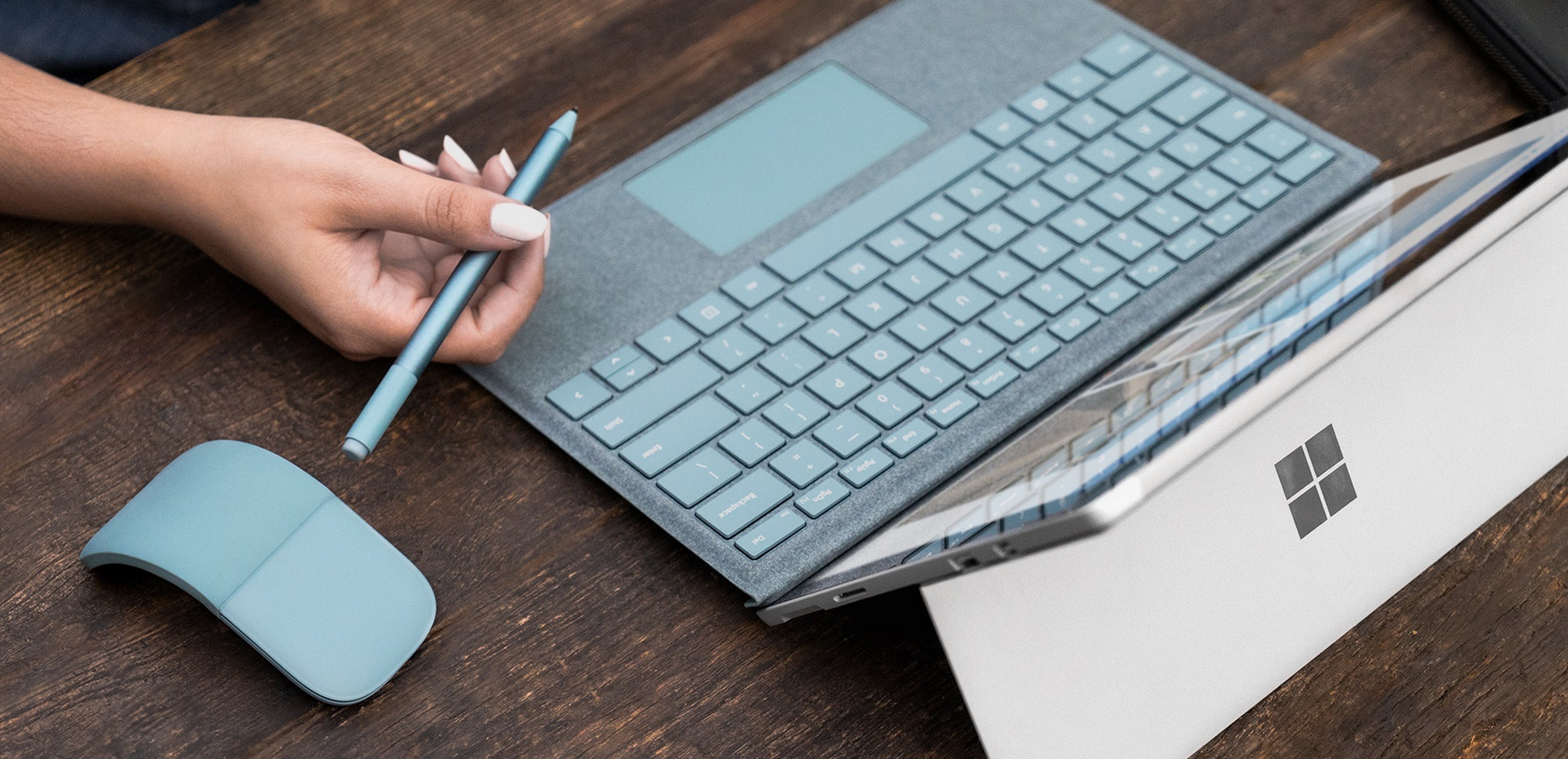 Person sitting at a table holding the new aqua Type Cover, Surface Arc Mouse, and Surface Pen.