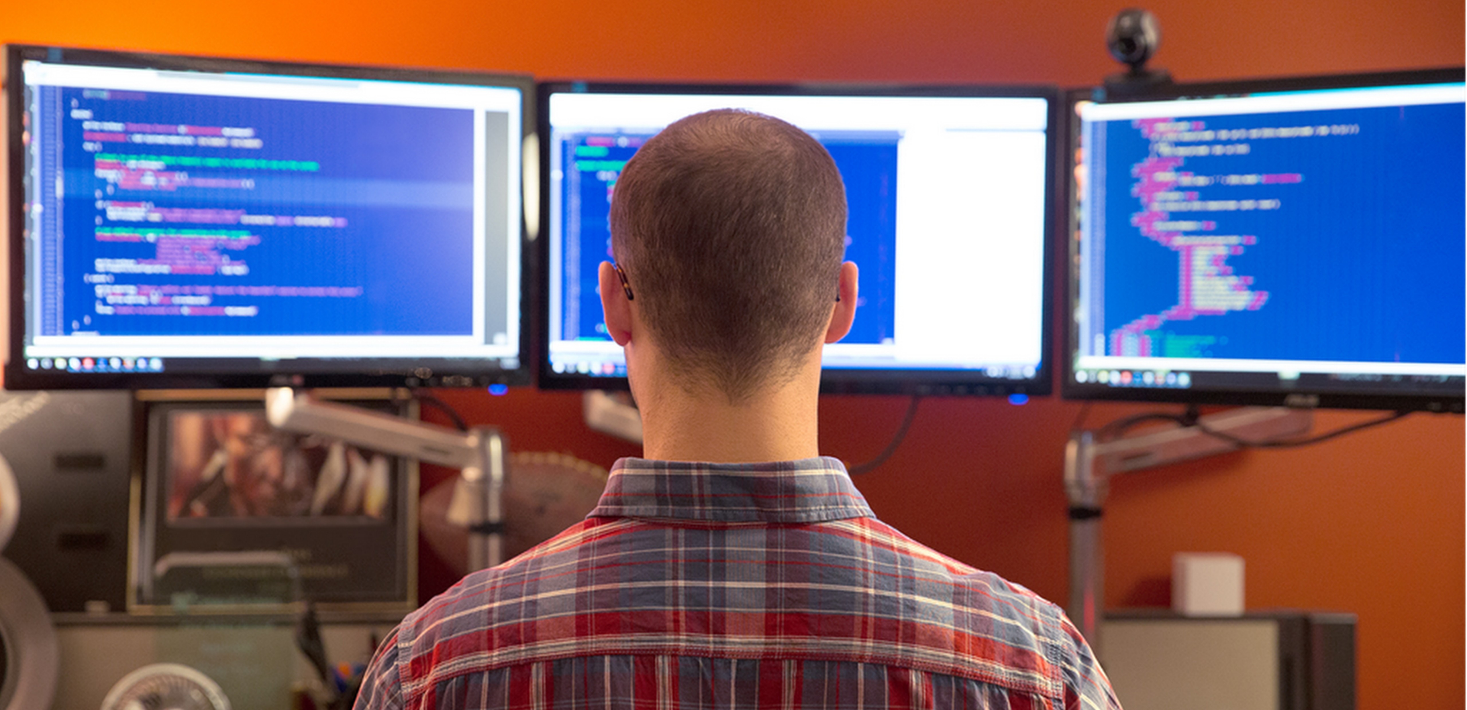 Man in front of three monitors in an office, the back of his head is shown