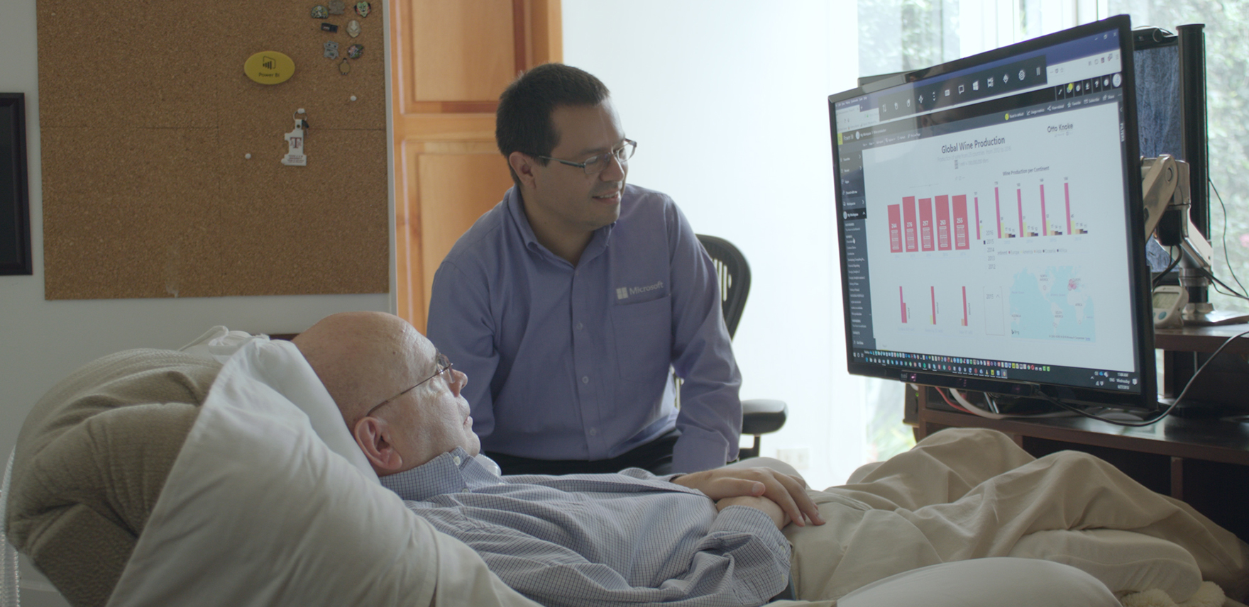 Older man (Otto Knoke) in a hospital bed and younger man (his friend and former colleague, Juan Alvarado) sitting beside him a Power BI report Knoke created using eye-tracking software for Windows 10 on a display monitor at the foot of the bed