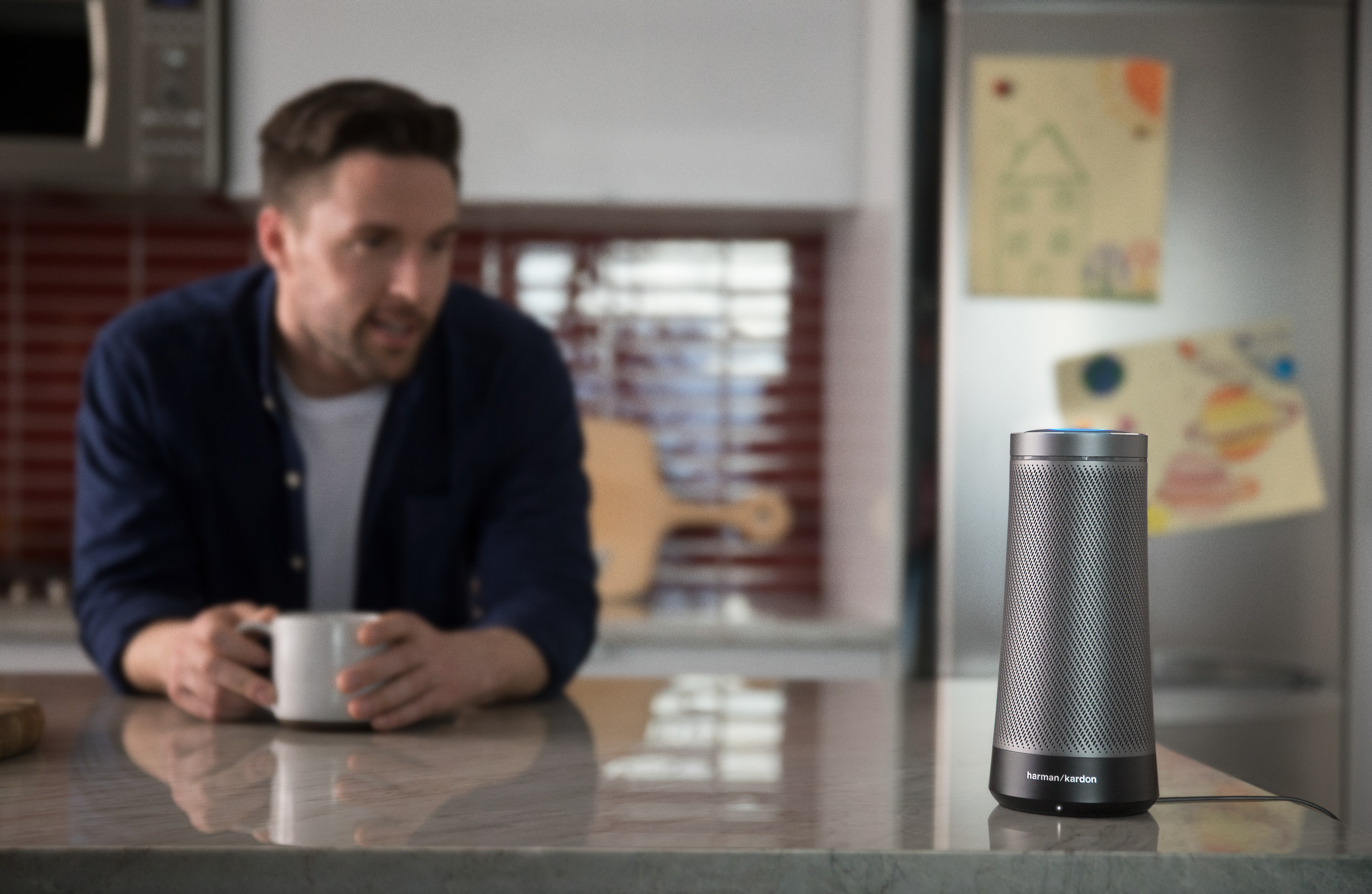 Man standing in kitchen, holding a cup of coffee looking at the Harman Kardon Invoke speaker that combines the rich, captivating sound that Harman Kardon is known for with your personal digital assistant, Cortana, in an innovative and beautifully designed speaker for the home.