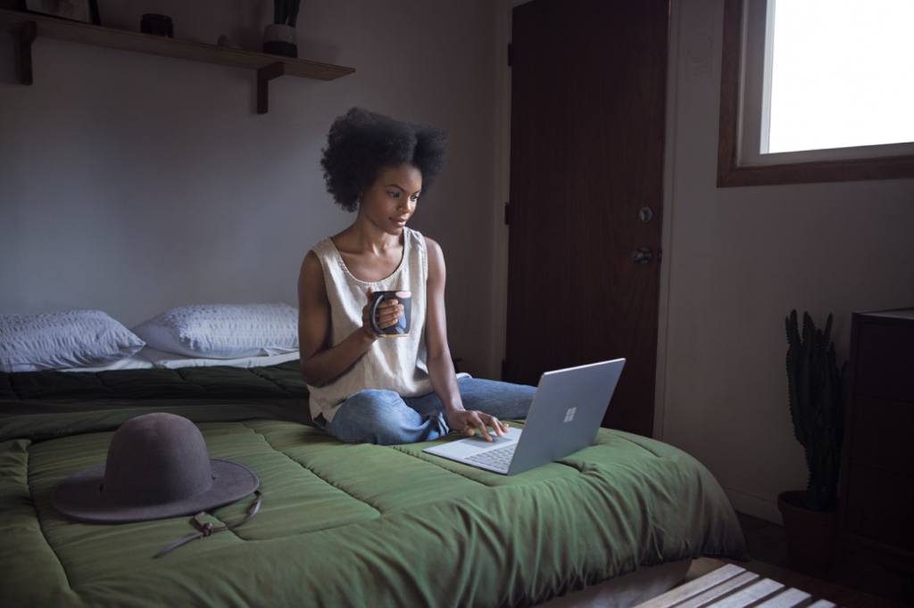 Woman sitting on bed holding a mug working on Surface Book.