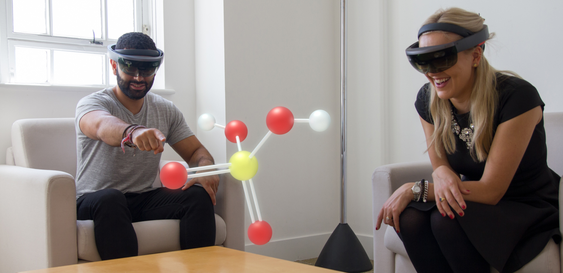 Man and woman sitting in a room wearing HoloLens and looking at a Hologram.