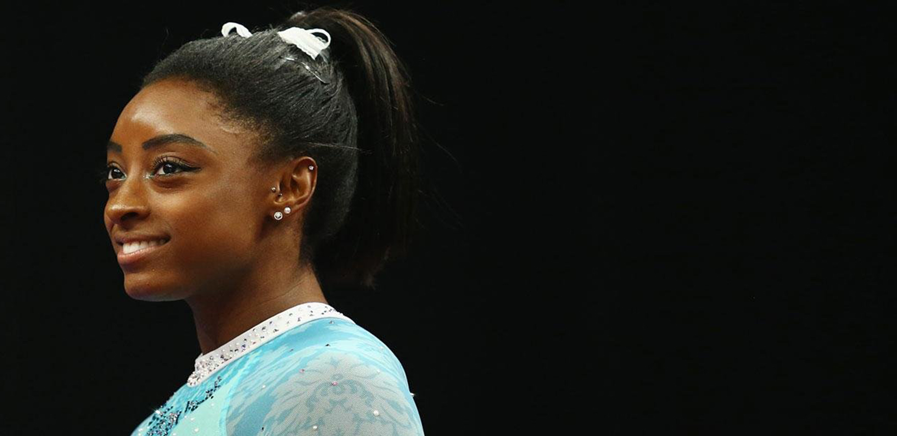 Simone Biles, standing and smiling, facing left, wearing a teal leotard, which is the color of sexual assault awareness.