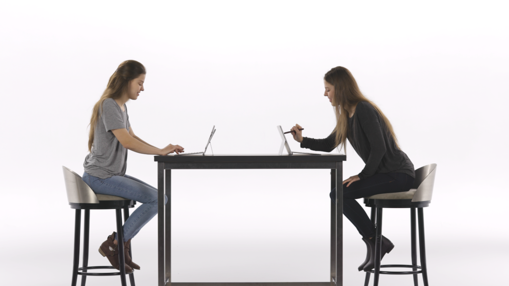 Two women, twins, sit across from each other at a table in front of PCs, with one using a digital pen to edit on the screen