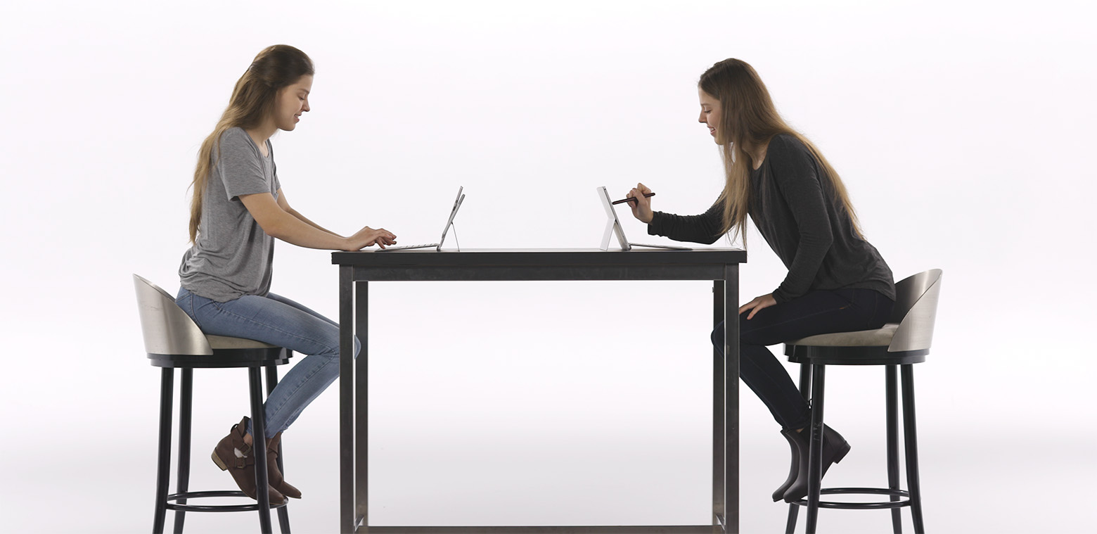 Two women, twins, sit across from each other at a table, in front of PCs, with one using a digital pen to work on the screen