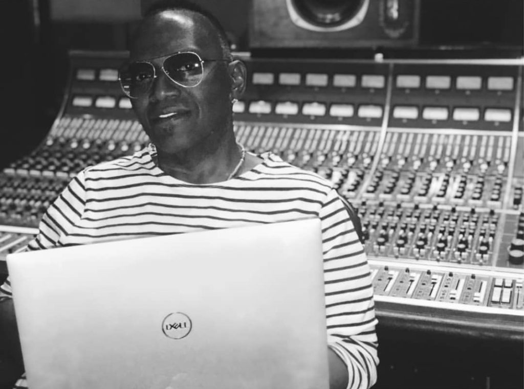 A black and white photo of Randy Jackson with studio equipment in the background, working on a Dell laptop