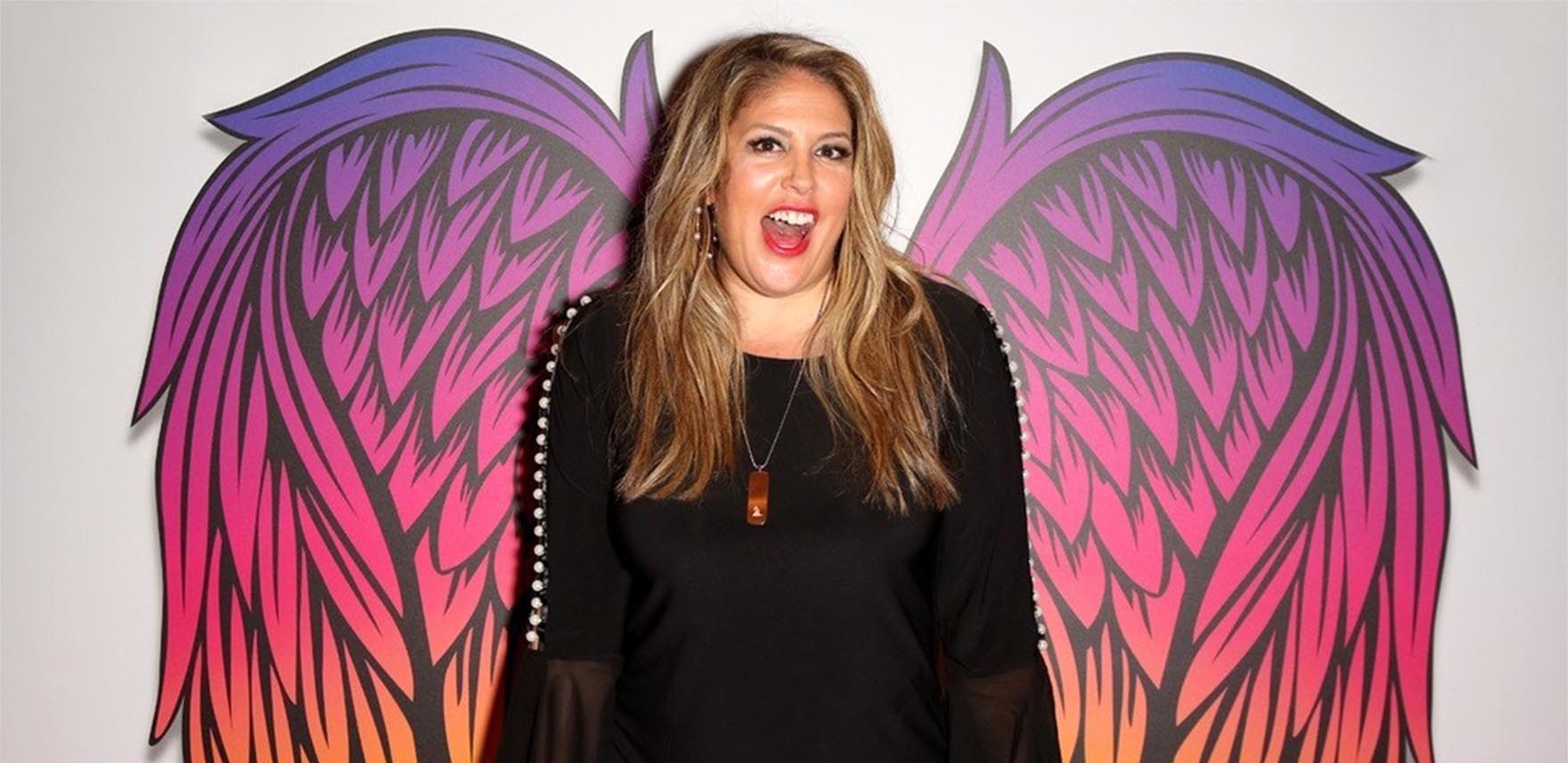 Lizza Monet Morales smiling in a black top with colorful wings behind her on a wall