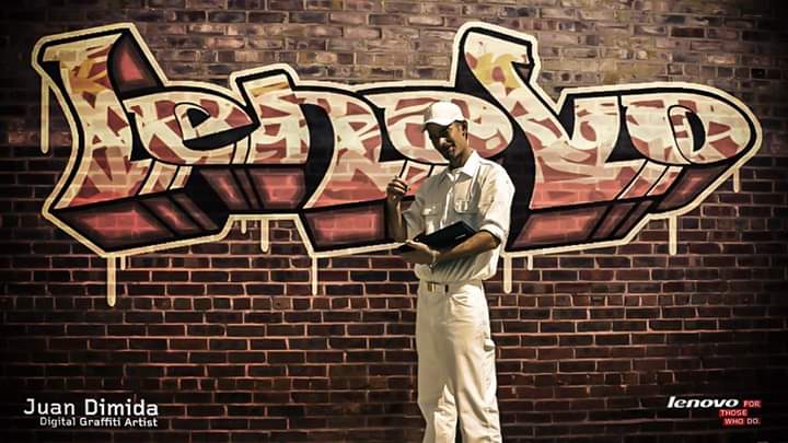 Juan Dimida standing with a Lenovo device in front of a brick wall with a graffiti-style Lenovo logo he drew