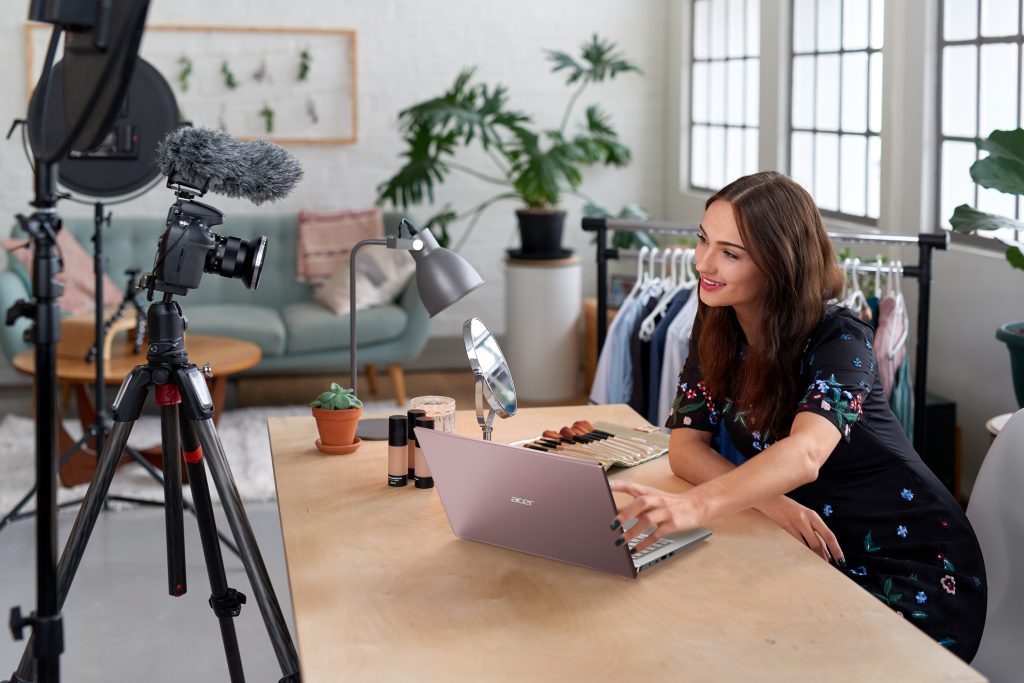 Woman sitting on a table in front of Acer Swift X while professional camera on a tripod aimed at her on a table