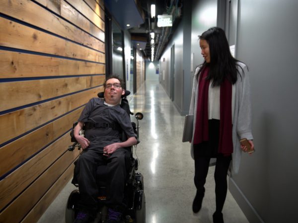 Man in a wheelchair travelling down a corridor with a woman