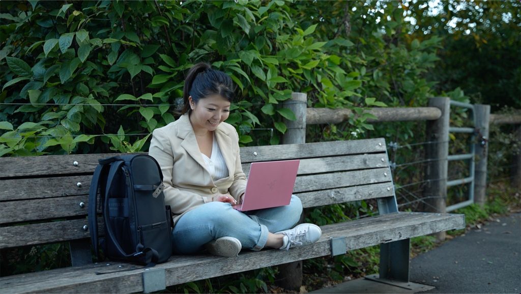 A woman sits on a bench outside while using a laptop
