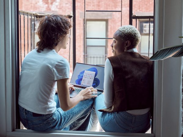 Two people talking while sitting in a window frame, one holding a laptop computer