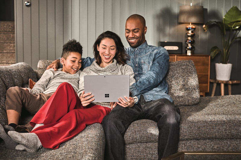 Family of three -- man, woman and boy -- sitting on a couch looking at a Surface device