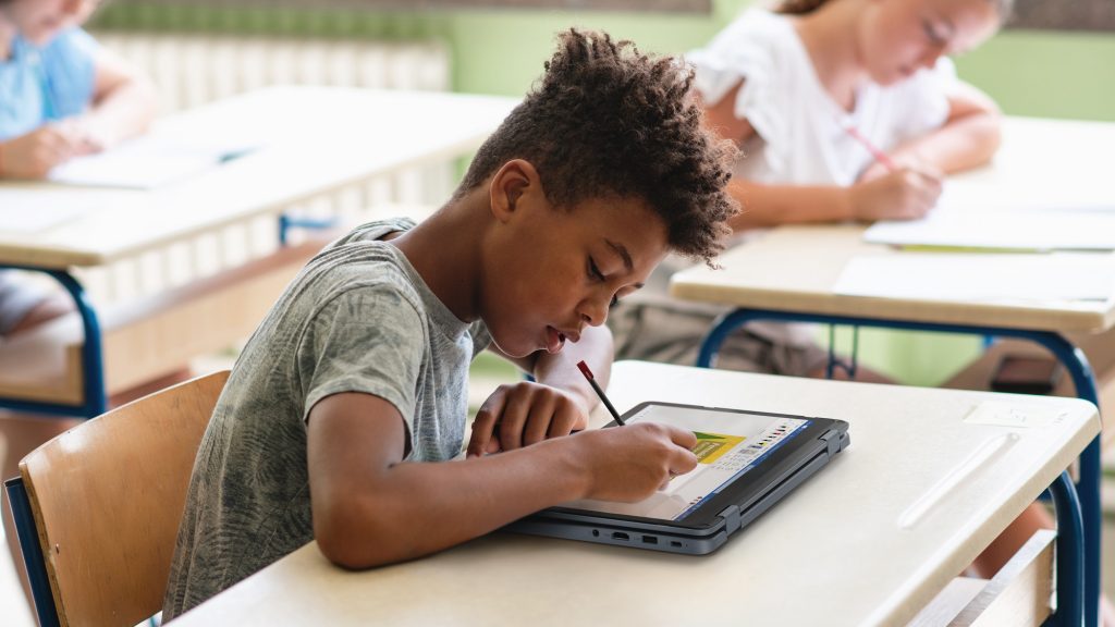 A young student uses a stylus on a laptop screen flat on his desk in a classroom
