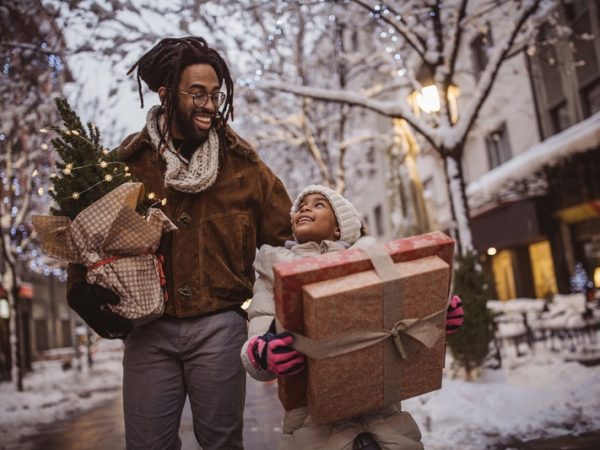 Father and child carrying gifts and a Christmas tree as they walk down a city street dusted with snow