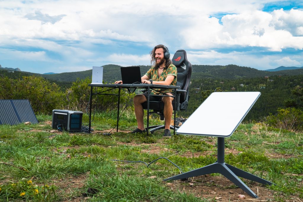 Man sitting on top of a mountain at a table with a laptop, satellite station nearby
