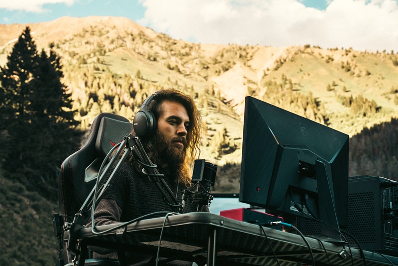 Man sitting at table wearing headphones and looking at PC display with mountains in background