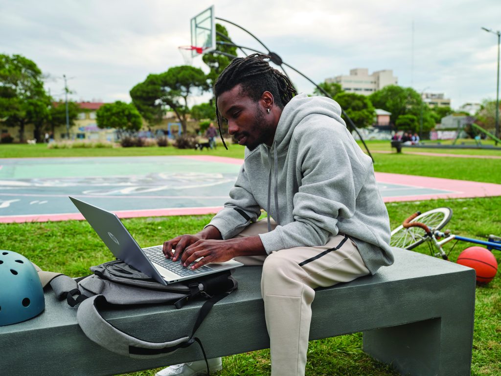 Young man working on a laptop computer next to an outdoors basketball court