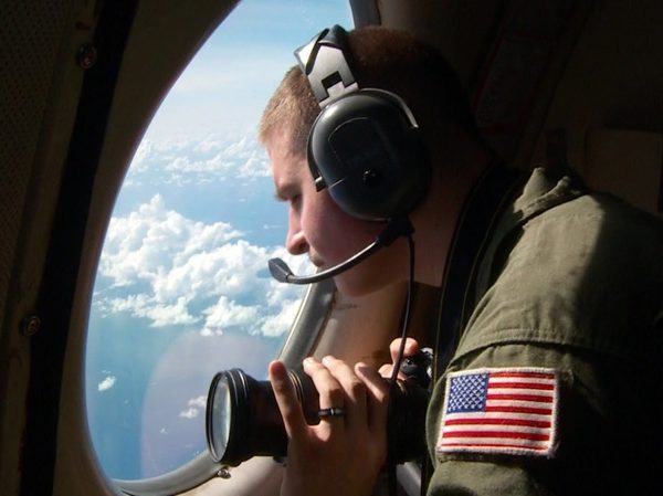 Man in uniform looking out large airplane window, holding a camera