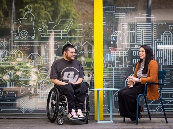 Man in a wheelchair conversing with a seated woman in an office lobby where walls are decorated with urban and rural scenes