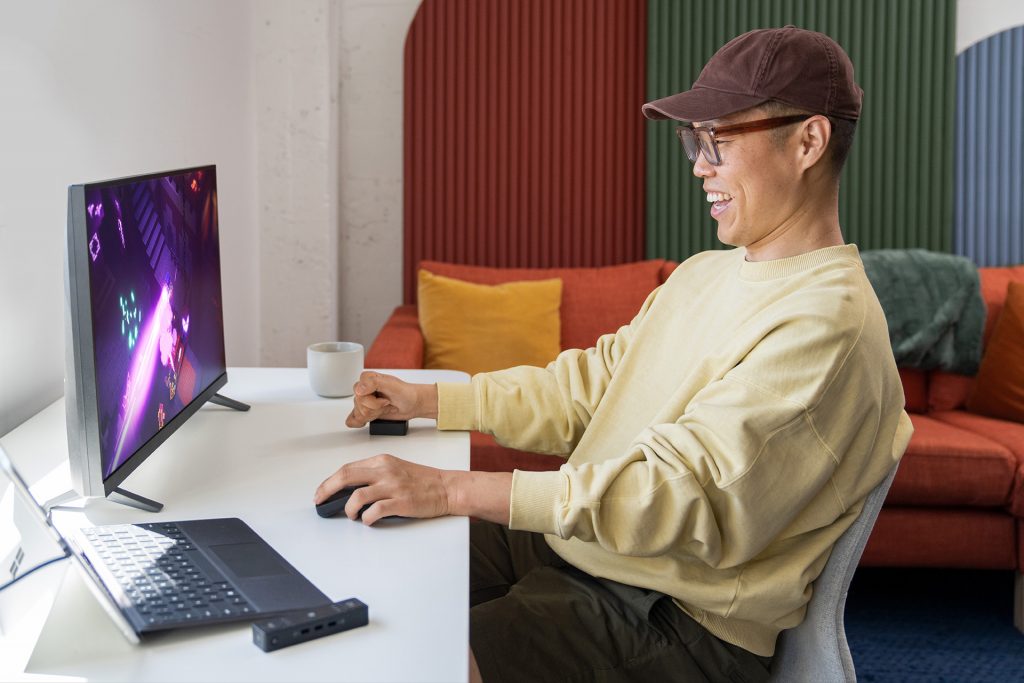 A man with a yellow sweater is gaming while using the Microsoft adaptive accessories. The man is sitting on a chair in front of a monitor that shows a racing game while using the Adaptive mouse with his left hand and the adaptive button with his right hand.