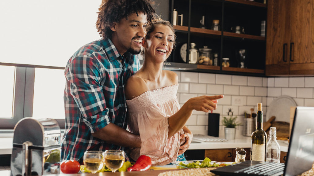 Foto: blackCAT/E+/Getty Images. Una mujer y un hombre sonríen mientras miran una receta en una laptop Windows y cocinan juntos